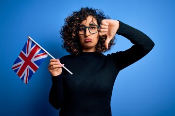 Sticker - Young beautiful curly arab woman wearing glasses holding uk flag over blue background with angry face, negative sign showing dislike with thumbs down, rejection concept