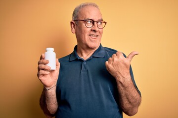 Wall Mural - Middle age hoary man holding medicine pills standing over isolated yellow background pointing and showing with thumb up to the side with happy face smiling