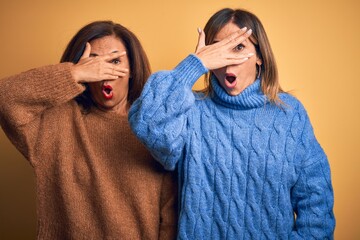 Poster - Middle age beautiful couple of sisters wearing casual sweater over isolated yellow background peeking in shock covering face and eyes with hand, looking through fingers with embarrassed expression.