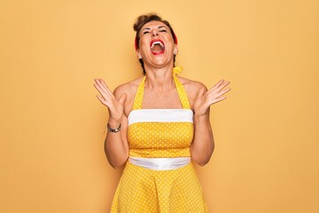 Sticker - Middle age senior pin up woman wearing 50s style retro dress over yellow background celebrating mad and crazy for success with arms raised and closed eyes screaming excited. Winner concept