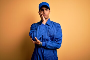Wall Mural - Young african american mechanic man wearing blue uniform and cap over yellow background thinking looking tired and bored with depression problems with crossed arms.