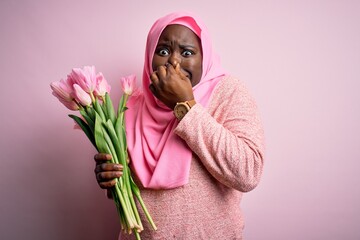 Poster - Young african american plus size woman wearing muslim hijab holding bouquet of pink tulips smelling something stinky and disgusting, intolerable smell, holding breath with fingers on nose. Bad smell