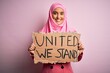 Young activist woman wearing pink muslim hijab holding banner with united we stand message with a happy face standing and smiling with a confident smile showing teeth