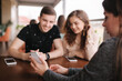© Aleksandr - Group of tree friends on terrace. Yound people meeting in cafe. Friends sitting in cafe together. Female show to couple something on her smartphone