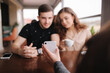 © Aleksandr - Group of tree friends on terrace. Yound people meeting in cafe. Friends sitting in cafe together. Female show to couple something on her smartphone