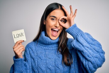 Wall Mural - Young beautiful brunette romantic woman holding reminder paper with love message with happy face smiling doing ok sign with hand on eye looking through fingers