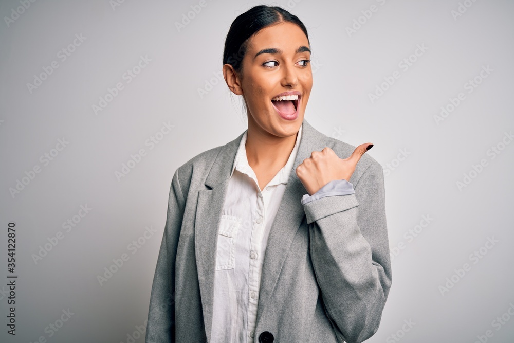 Young beautiful brunette businesswoman wearing jacket standing over white background smiling with happy face looking and pointing to the side with thumb up.