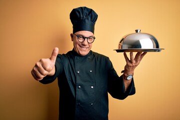 Poster - Middle age handsome grey-haired waiter man wearing cooker uniform and hat holding tray approving doing positive gesture with hand, thumbs up smiling and happy for success. Winner gesture.