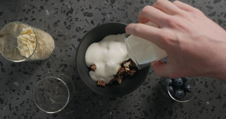 top view man hands preparing breakfast with chocolate granola, almond flakes and blueberry in black bowl on terrazzo surface