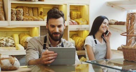 Caucasian man vendor using tablet device while standing at the counter in the bakery, his female co-worker talking on the phone behind him. Inside