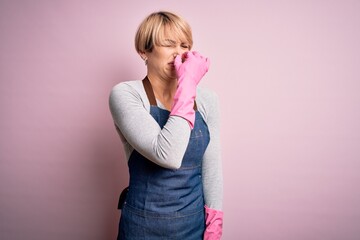 Wall Mural - Young blonde cleaner woman with short hair wearing apron and gloves over pink background smelling something stinky and disgusting, intolerable smell, holding breath with fingers on nose. Bad smell