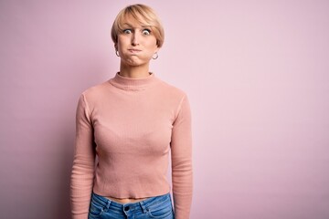 Poster - Young blonde woman with short hair wearing casual turtleneck sweater over pink background puffing cheeks with funny face. Mouth inflated with air, crazy expression.