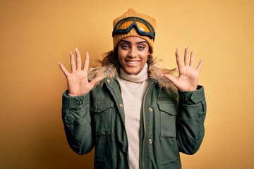 Wall Mural - Young african american afro skier girl wearing snow sportswear and ski goggles showing and pointing up with fingers number ten while smiling confident and happy.