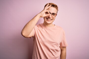 Wall Mural - Young handsome redhead man wearing casual t-shirt standing over isolated pink background doing ok gesture with hand smiling, eye looking through fingers with happy face.