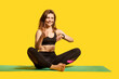 © khosrork - Joyful sporty girl with fitness tracker on her wrist, sitting on mat and showing thumbs up, like gesture, satisfied with pulse, heart beat indicators in smartwatch after sport, wearable app. Health