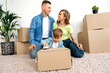 Relocation. The young family moved into their new home. They are sitting on the floor in the middle of cardboard boxes with things in a happy mood. Their little cute son is playing with a toy car