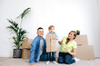 Relocation. The young family moved into their new home. They are sitting on the floor in the middle of cardboard boxes in a happy mood and their little cute son holds a cardboard box and smiling