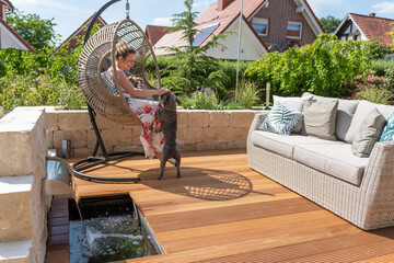 Attractive blonde woman and dog relaxing on a wooden terrace with green garden and modern outdoor furniture on a sunny day.
