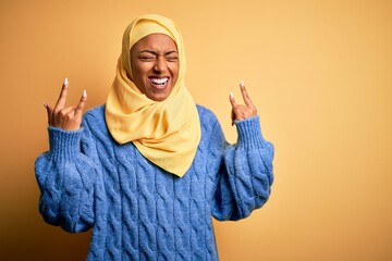 Wall Mural - Young African American afro woman wearing muslim hijab over isolated yellow background shouting with crazy expression doing rock symbol with hands up. Music star. Heavy concept.