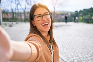 Young beautiful brunette woman wearing glasses smiling happy and confident. Standing with smile on face making selfie by the camera at street of city