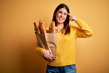 Poster - Young beautiful woman holding a bag of fresh healthy bread over yellow background Smiling pointing to head with one finger, great idea or thought, good memory