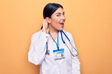 Sticker - Young beautiful doctor woman wearing stethoscope and id card over isolated yellow background smiling with hand over ear listening and hearing to rumor or gossip. Deafness concept.