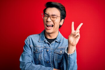 Wall Mural - Young handsome chinese man wearing denim jacket and glasses over red background smiling with happy face winking at the camera doing victory sign. Number two.