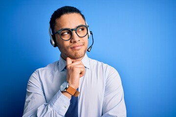 Young brazilian call center agent man wearing glasses and tie working using headset with hand on chin thinking about question, pensive expression. Smiling with thoughtful face. Doubt concept.