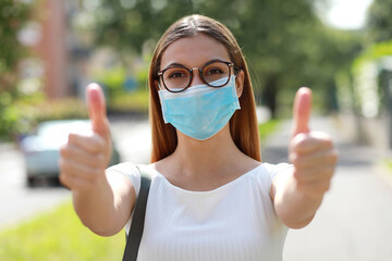 Wall Mural - Portrait of optimistic girl wearing protective mask showing thumbs up in city street