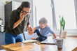 © Vadim Pastuh - A toddler boy interferes mother with work at home. A young mom in eyeglasses watching on laptop and writes in notebook while kid sits near on a table