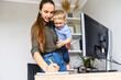 © Vadim Pastuh - A young mother in casual wear working remotely, woman with a toddler on the arms writes notes in planner on a pc desk in home office