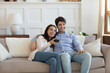 © fizkes - Overjoyed young caucasian couple sit relax on comfortable sofa in living room watching TV together, happy millennial man and woman rest on couch at home enjoy television program on leisure weekend