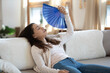 © fizkes - Exhausted young caucasian woman sit relax on couch in living room wave with hand fan suffer from hot weather, overheated female use waver feel unwell unhealthy lack air condition system indoors