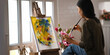 © Prathankarnpap - Woman painting on canvas with paintbrush while sitting at the chair over modern and comfortable art studio windows as background.  Concept of working artist woman.