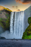 Two male and female tourists walking and watching the amazing waterfalls of Skogafoss in southern Iceland. In the morning, the sun rises from behind the mountains with green grass all over the area.