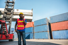 Black Foreman Worker Working Control The Crane And Forklift At Container Cargo Harbor To Loading Containers. African Dock Male Staff With Radio For Logistics Import Export Shipping Concept.