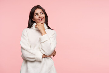 Wall Mural - Studio photo of young pretty brunette woman with loose hair touching her face with raised hand while looking dreamiliy upwards, standing over pink background