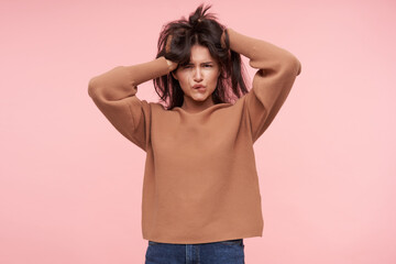 Confused young attractive brown haired woman lady rumpling her hair and squinting eyes while looking at camera, posing over pink background in casual wear