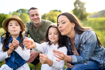 Happy family with two small daughters sitting outdoors in spring nature, blowing dandelion seeds.