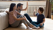 © fizkes - Happy young parents sit on warm wooden floor play have fun with excited preschooler son, smiling family relax rest in cozy home living room, engaged in funny game activity with little kid