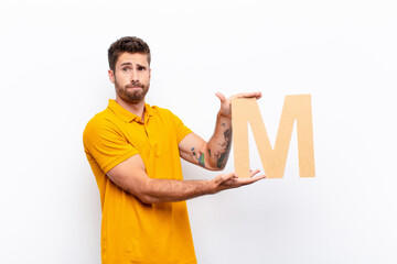 young handsome man confused, doubtful, thinking, holding the letter M of the alphabet to form a word or a sentence.