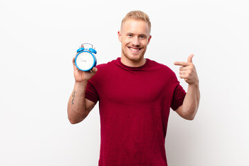 Wall Mural - young blonde man smiling confidently pointing to own broad smile, positive, relaxed, satisfied attitude against white wall holding an alarm clock