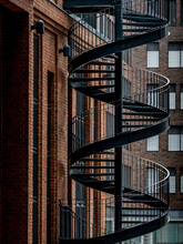Frontal View Of A Steel Spiral Staircase In Industrial Design Leading Upwards Next To A Brick Facade In A Modern Backyard Outside Of An Office Building