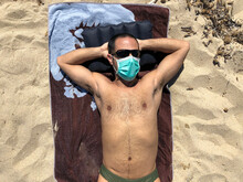Ragazzo Moro Con Occhiali Prende Il Sole Disteso Sull'asciugamano In Spiaggia Indossando Una Mascherina Chirurgica