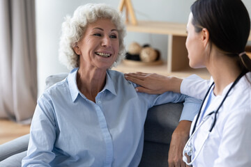 Wall Mural - Aged woman talks with young caregiver seated on couch at home, female nurse in white coat puts hand on patient shoulder showing care support and friendly warm relation at visit homecare of old person