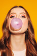 young adult girl inflates pink bubble gum isolated on yellow background, close up portrait