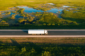 Wall Mural - Semi truck with white freight trailer drive on highway in green grass countryside at sunrise, aerial top view. Car with motion blur effect. Road transportation and logistics background