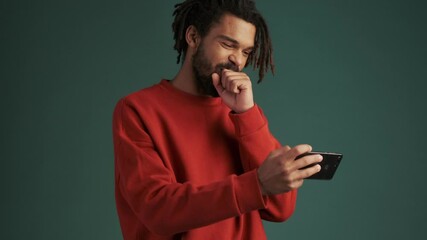 Wall Mural - A happy young african american man is watching something interesting on his smartphone standing isolated over green wall background in studio