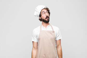 Wall Mural - young crazy chef with a worried, confused, clueless expression, looking up to copy space, doubting against white wall