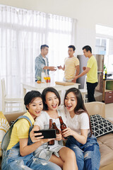 Canvas Print - Pretty young Vietnamese women clinking beer bottles and talking selfie at house party